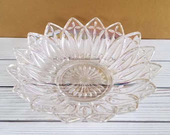 May include: A clear glass bowl with a scalloped edge and a starburst pattern in the center. The bowl is sitting on a white wooden surface.