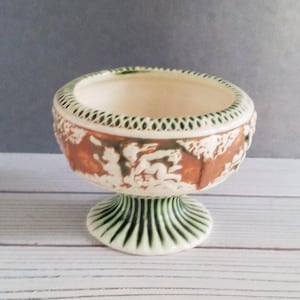 May include: A pedestal bowl with a cream-colored interior and a detailed exterior. The bowl features a brown and white relief design around the sides and a green rim. The base is green with vertical lines.