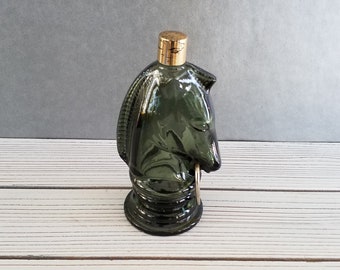 May include: A dark green glass bottle shaped like a knight's head with a gold cap. The bottle is sitting on a white wooden surface.