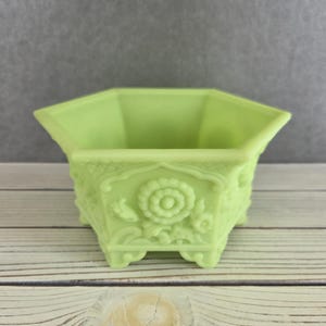 May include: A light green, hexagonal planter with ornate floral and leaf detailing. The planter has a square base and is made of a solid, opaque material. The background is a neutral gray, and the planter sits on a light-colored wooden surface.