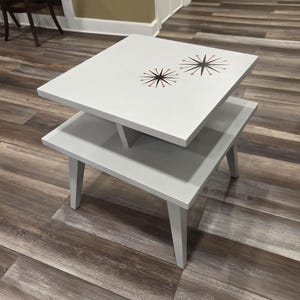 May include: A two-tiered, square side table painted light gray. The top tier features two starburst designs in black, orange, and brown. The table has four legs and a lower shelf. The table is approximately 24 inches wide and 24 inches long.
