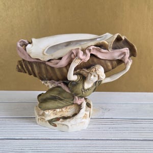 May include: A decorative ceramic sculpture featuring a woman figure supporting a large seashell. The sculpture is painted in shades of green, pink, and cream, with a gold background. The seashell is detailed with textured edges.