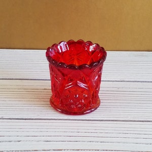 May include: A small, red glass toothpick holder with a decorative, diamond-shaped pattern. The holder has a scalloped rim and a clear base.