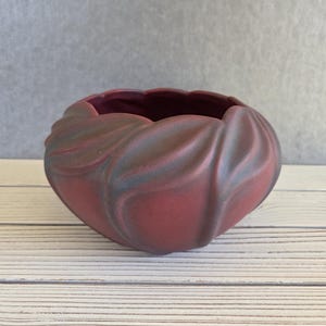 May include: A maroon-colored ceramic bowl with a textured, petal-like design. The bowl has a wide opening and sits on a white wooden surface. The bowl's exterior has a matte finish with subtle color variations.