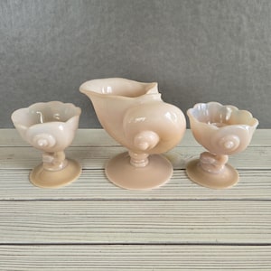 May include: Three light peach-colored glass serving pieces. The set includes a tall, ornate pitcher and two smaller stemmed bowls. The pieces have a seashell-like design and sit on a light-colored wooden surface.