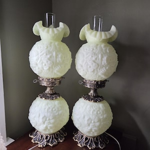 May include: A pair of matching table lamps featuring pale yellow, translucent glass globes with embossed floral patterns. Each lamp has a ruffled glass top, a clear glass chimney, and a dark metallic base with ornate, leaf-like detailing.