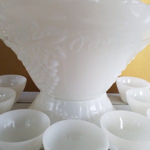 May include: A white milk glass punch bowl with a grapevine design and matching cups. The bowl has a scalloped edge and the cups have a small handle.