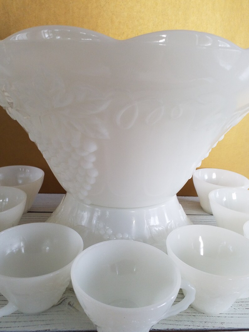 May include: A white milk glass punch bowl with a grapevine design and matching cups. The bowl has a scalloped edge and the cups have a small handle.
