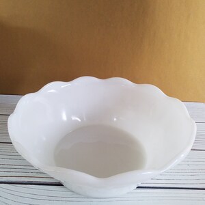 May include: A white, scalloped-edge bowl made of glass. The bowl is empty and sitting on a white wooden surface.