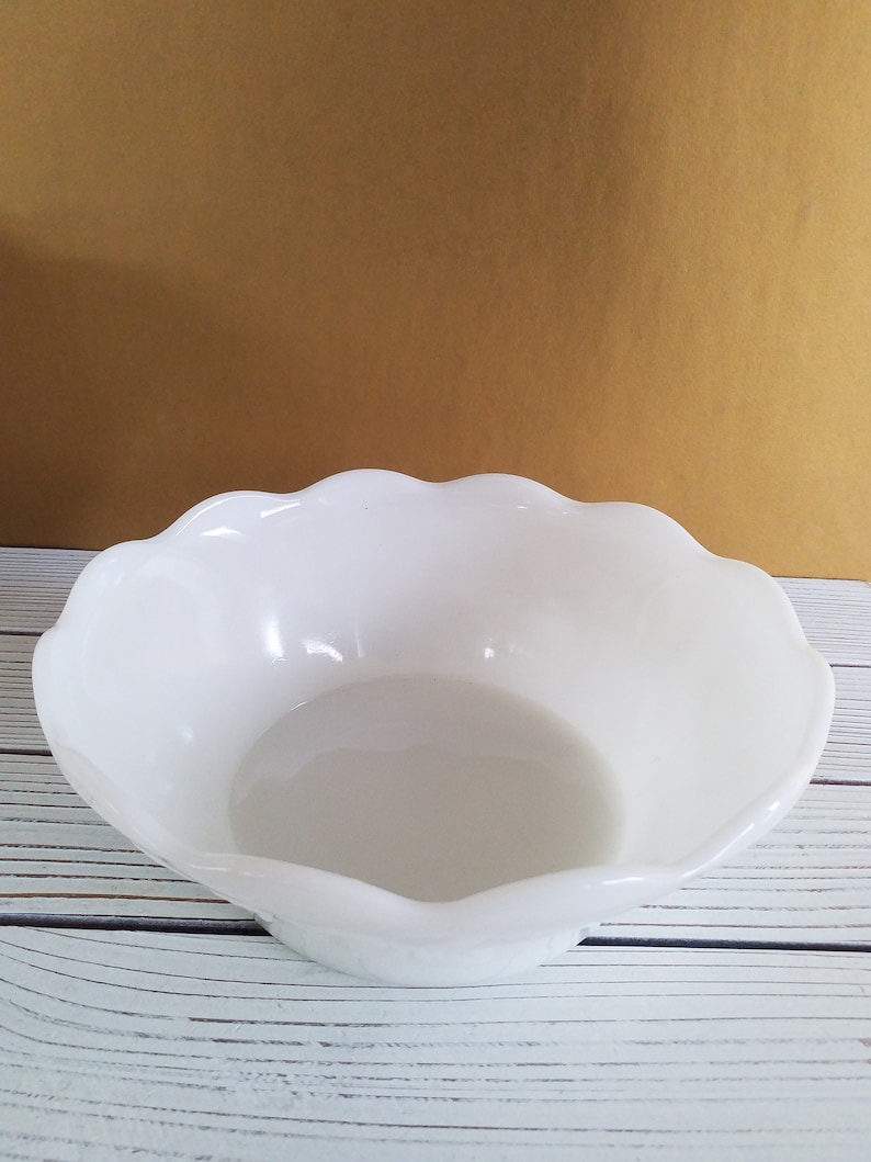 May include: A white, scalloped-edge bowl made of glass. The bowl is empty and sitting on a white wooden surface.