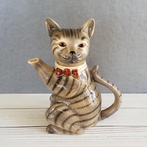 May include: A ceramic teapot shaped like a gray tabby cat wearing a red bow tie. The cat has a white collar and is sitting on a white surface.