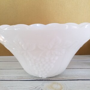 May include: White milk glass punch bowl with a grape design. The bowl has a scalloped edge and is sitting on a whitewashed wooden surface.