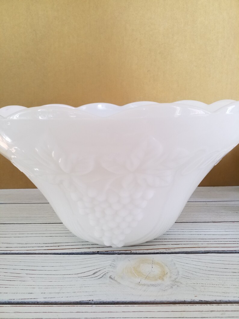 May include: White milk glass punch bowl with a grape design. The bowl has a scalloped edge and is sitting on a whitewashed wooden surface.