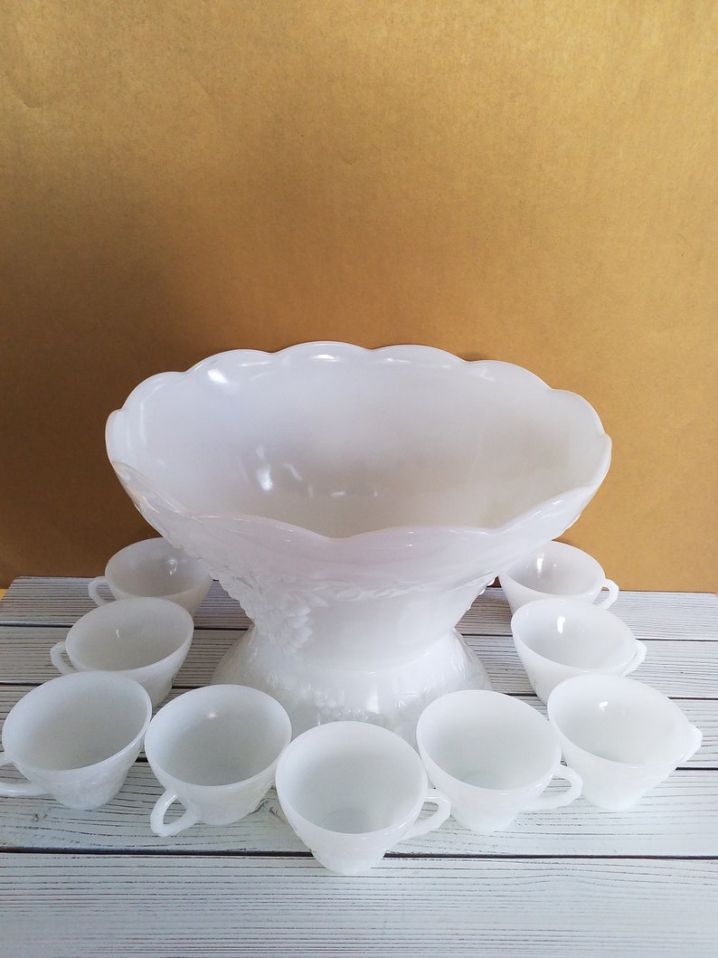 May include: A white milk glass punch bowl with a scalloped edge and a grapevine design. The bowl is surrounded by 10 matching white milk glass cups.