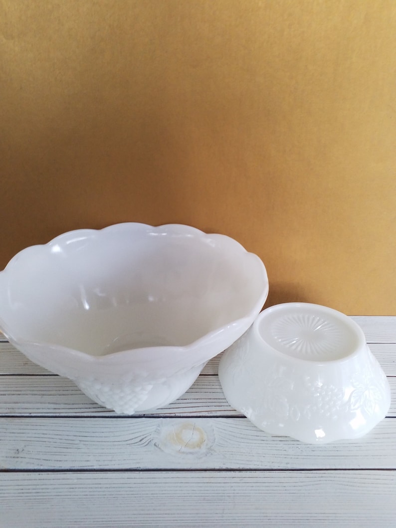 May include: Two white milk glass bowls with a grape design. The larger bowl is upside down and the smaller bowl is right side up.