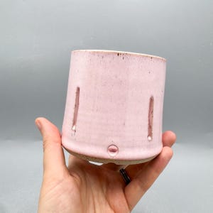 May include: A cylindrical ceramic planter with a light pink glaze and a speckled texture. The vessel features vertical cutouts along the sides and a small circular drainage hole near the base, which has an exposed, unglazed tan clay rim.