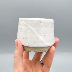 May include: A set of three ceramic mugs with a speckled, matte finish. The mugs are in shades of cream, light gray, and dark gray. Each mug has a simple, minimalist design and a rounded handle.