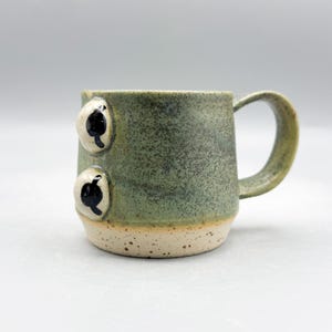 May include: A handmade ceramic mug with a green and tan speckled glaze. The mug features two applied, rounded, white and black eye-shaped designs on the side. The handle is a matching green color.