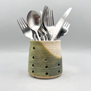 Puede incluir: Un recipiente de cerámica para utensilios en verde y crema, lleno de cubiertos de acero inoxidable. El recipiente tiene agujeros circulares y un acabado moteado. Los cubiertos incluyen tenedores, cucharas y cuchillos.