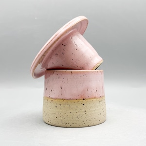 Puede incluir: Un plato de mantequilla de cerámica de dos piezas con tapa. La tapa y la parte superior de la base son de color rosa moteado, mientras que la parte inferior es de color beige moteado. La tapa descansa sobre la base.