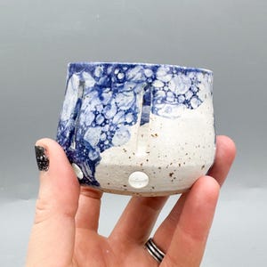 Puede incluir: Una taza de cerámica hecha a mano con un diseño único. La taza presenta una base blanca con detalles moteados y un patrón azul en forma de burbujas en la mitad superior. Tiene secciones recortadas y un agujero redondo cerca de la base.