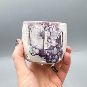 Puede incluir: Una maceta de cerámica hecha a mano con base blanca y diseño morado veteado. La maceta tiene formas rectangulares recortadas y pequeños orificios de drenaje. El borde es de un tono morado más oscuro.