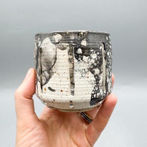 Peut inclure: Une tasse en céramique faite à la main avec un design unique. La tasse présente une base blanche mouchetée, avec des motifs abstraits noirs et gris et des lignes verticales découpées. Le bord est gris foncé. La tasse est tenue dans une main.