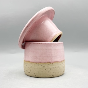 May include: A ceramic butter keeper featuring a two-tone design with a glossy light pink glaze on the upper portion and a speckled cream-coloured base. The lid is tilted and rests atop the cylindrical container, showcasing a smooth finish.