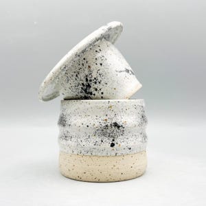 Op de afbeelding: Twee handgemaakte keramische stukken. Het onderste stuk is een witte beker met een beige basis en een gestructureerd, geribbeld ontwerp. Het bovenste stuk is een wit, kegelvormig deksel met zwarte spikkels. De stukken zijn op elkaar gestapeld.