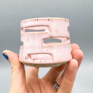 以下が含まれることがあります： 淡いピンク色の小さな円筒形のセラミックキャンドルホルダー。ホルダーの本体には長方形の切り抜きが施されています。ベースはまだらの自然な仕上がりです。キャンドルホルダーは手で持たれています。