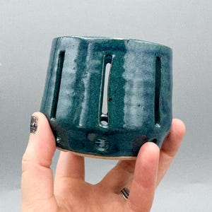 Peut inclure: Un récipient en céramique turquoise avec des fentes verticales et de petites indentations circulaires. La poterie faite à la main a une finition brillante et est tenue dans une main sur un fond neutre. La forme du récipient est légèrement conique.