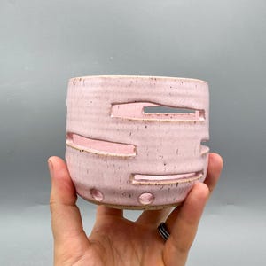 Peut inclure: Un récipient cylindrique en céramique avec un émail rose clair moucheté, présentant des découpes horizontales et deux petites perforations circulaires près de la base. La surface extérieure possède une texture légèrement nervurée.