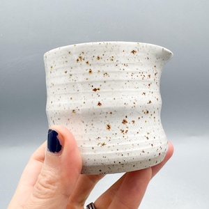 May include: A handmade ceramic creamer with a speckled white glaze and a small spout. The creamer has a textured, ridged design and is accented with brown speckles. The creamer is held in a hand against a gray background.