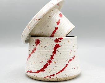 May include: A ceramic butter dish with a lid. The dish and lid are off-white with red splatter paint. The dish is cylindrical with a slightly flared base. The lid is round and sits on top of the dish.