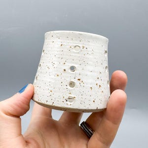 Puede incluir: Un portavelas votivo de cerámica blanca moteada con varias aberturas circulares. La cerámica hecha a mano tiene una forma ligeramente acampanada. El portavelas se sostiene en una mano sobre un fondo gris.