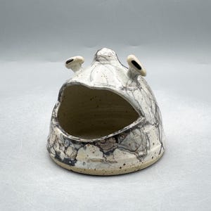 Peut inclure: Une sculpture en céramique avec une large bouche ouverte et deux petites protubérances arrondies. La pièce est principalement blanche avec un motif marbré de gris et de brun. L'intérieur est de couleur beige clair et la base est tachetée.