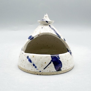 Peut inclure: Un salière en céramique faite à la main, avec une base blanche et un motif éclaboussé de bleu. Le salière a une forme triangulaire avec un petit couvercle pointu sur le dessus. L'intérieur est non émaillé, et la base est de couleur naturelle mouchetée.