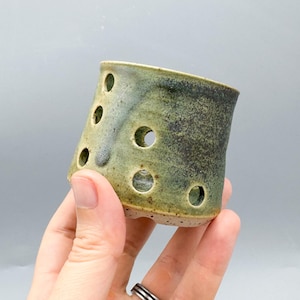 May include: A small, cylindrical ceramic vessel featuring a mottled green and brown glaze with a series of circular cutouts distributed across its surface. The base of the pottery piece shows a speckled, light-colored clay texture.