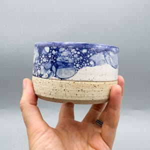 Puede incluir: Un cuenco cilíndrico de cerámica con una base beige moteada y una sección superior en contraste decorada con un patrón de burbujas azul y blanco. El recipiente tiene un borde liso y un acabado texturizado en la mitad inferior.