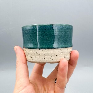 May include: A handmade ceramic cup with a teal glaze on the upper half and a speckled, unglazed beige base. The cup is cylindrical and appears to be held by a hand. The cup is approximately 3 inches in diameter and 2.5 inches tall.