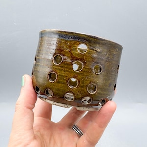 Può includere: Un vaso di ceramica fatto a mano con una glassa marrone scuro e verde oliva. Il vaso cilindrico presenta numerosi fori circolari sui lati. Il vaso è tenuto da una mano su uno sfondo grigio. Il fondo del vaso è di un colore più chiaro.