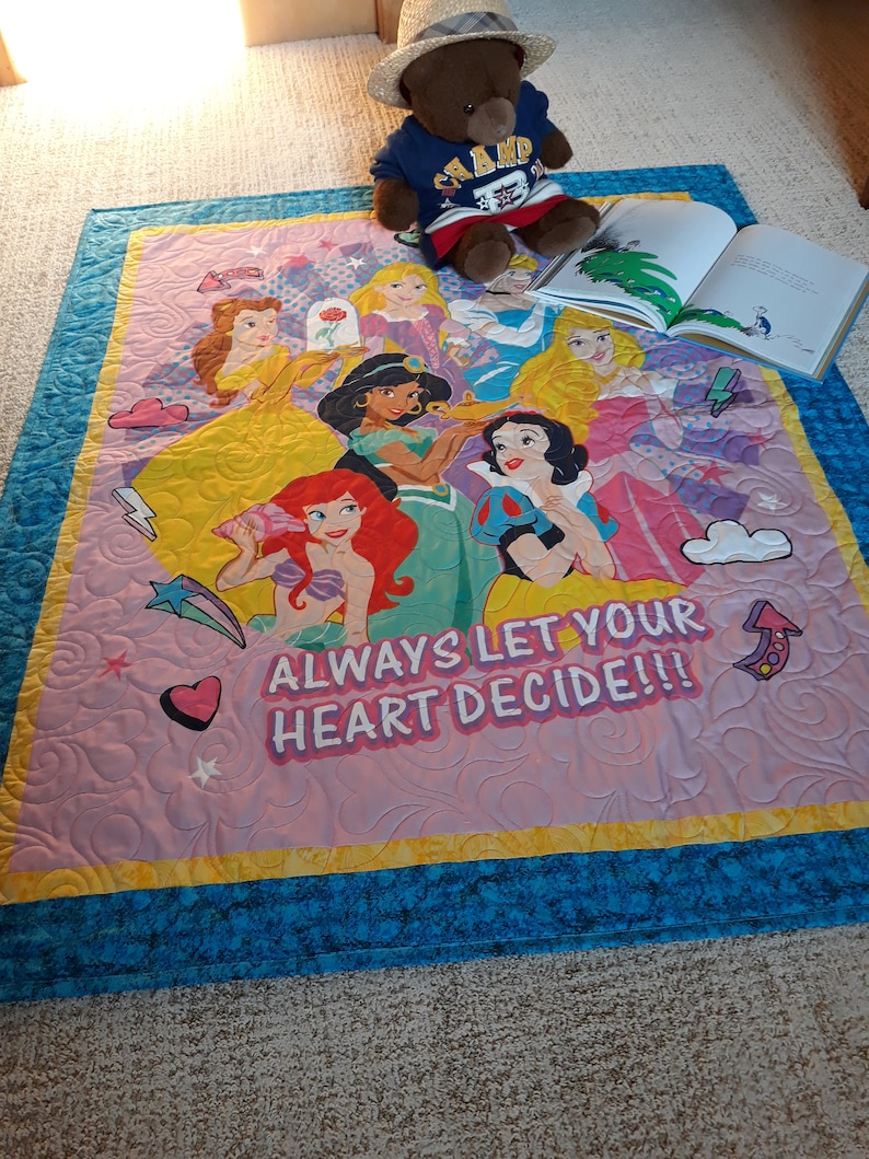 May include: A pink and blue quilted blanket with a colorful image of Disney princesses and the text "Always let your heart decide!!".