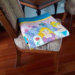 May include: A pink, blue, and yellow quilt with a princess theme. The quilt features two princesses with blonde hair and a rose with a green stem. The quilt is folded and placed on a wooden chair.