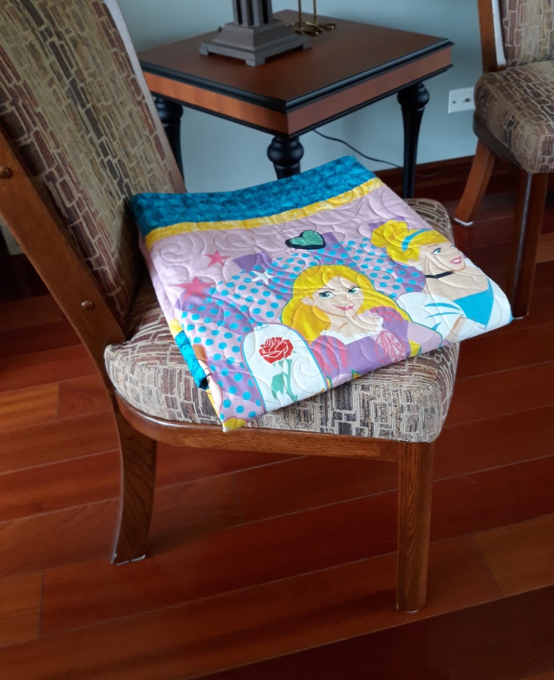 May include: A pink, blue, and yellow quilt with a princess theme. The quilt features two princesses with blonde hair and a rose with a green stem. The quilt is folded and placed on a wooden chair.