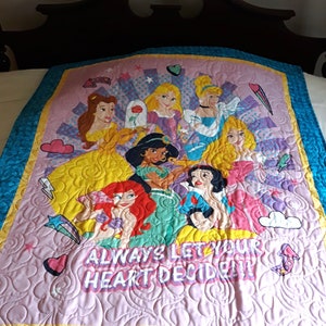 May include: A pink and yellow quilted throw blanket with a design featuring Disney princesses and the text "Always let your heart decide".