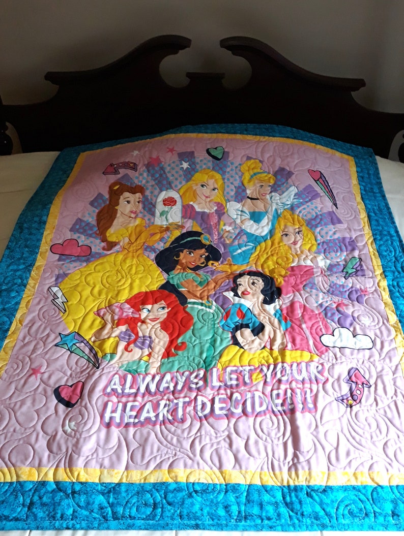 May include: A pink and yellow quilted throw blanket with a design featuring Disney princesses and the text "Always let your heart decide".