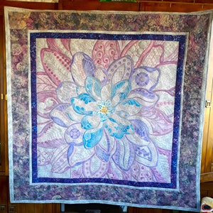 Könnte beinhalten: Ein gesteppter Wandbehang mit einem großen, filigranen Blumendesign in Violett-, Blau- und Weißtönen. Die Blume ist von kleineren Blumenmotiven umgeben und hat eine detaillierte, strukturierte Oberfläche.