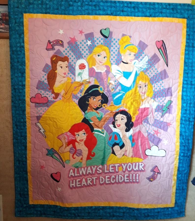 May include: A pink and purple quilt with a colorful design featuring Disney princesses. The princesses are surrounded by stars, hearts, and clouds. The text "ALWAYS LET YOUR HEART DECIDE!!!" is printed on the quilt.