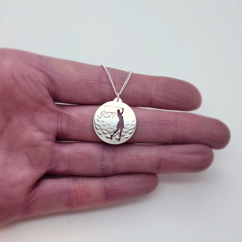 May include: Silver necklace with a round pendant featuring a golfer silhouette and a golf ball design. The pendant has the number "197" engraved on it.