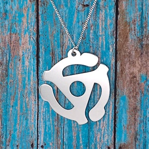 May include: A silver necklace with a pendant shaped like a vintage 45 rpm record adapter. The pendant is hanging against a blue wooden background.